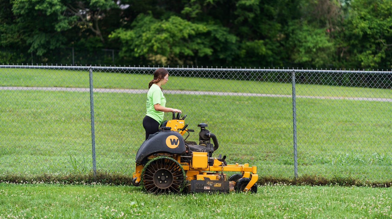 A commercial lawn care contractor with a riding mower looking out at a field next to a fence