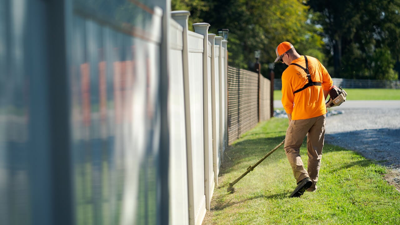 man using weed whacker to trim grass along fence