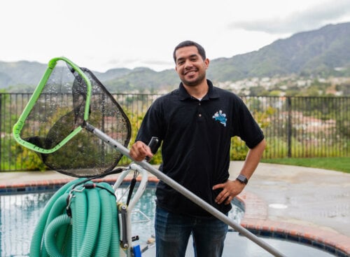 pool company names: pool cleaning employee standing in front of a pool holding skimmer