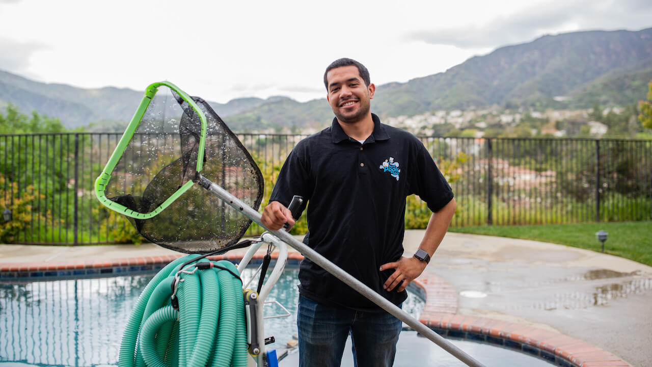 pool company names: pool cleaning employee standing in front of a pool holding skimmer