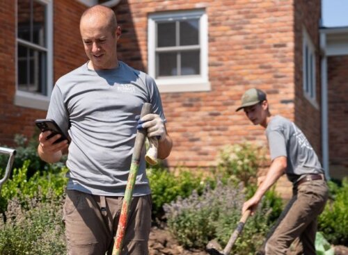 Two landscaping technicians on site, one digging and the other looking at their phone