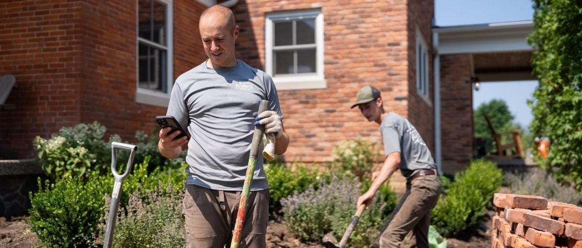 Two landscaping technicians on site, one digging and the other looking at their phone