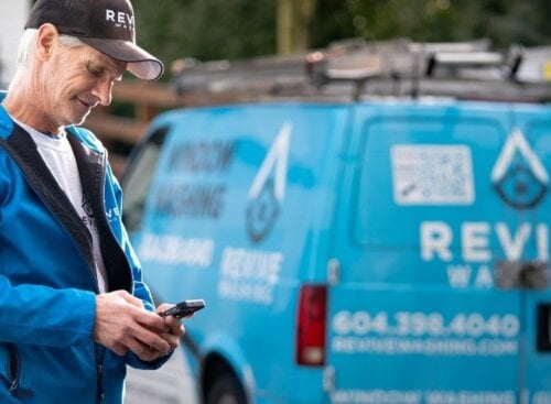 Window washing technician standing outside residential home looking down at their mobile device