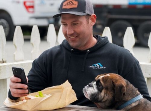 Roofing service provider sits at a table with a bulldog beside him, looking at his phone while eating lunch