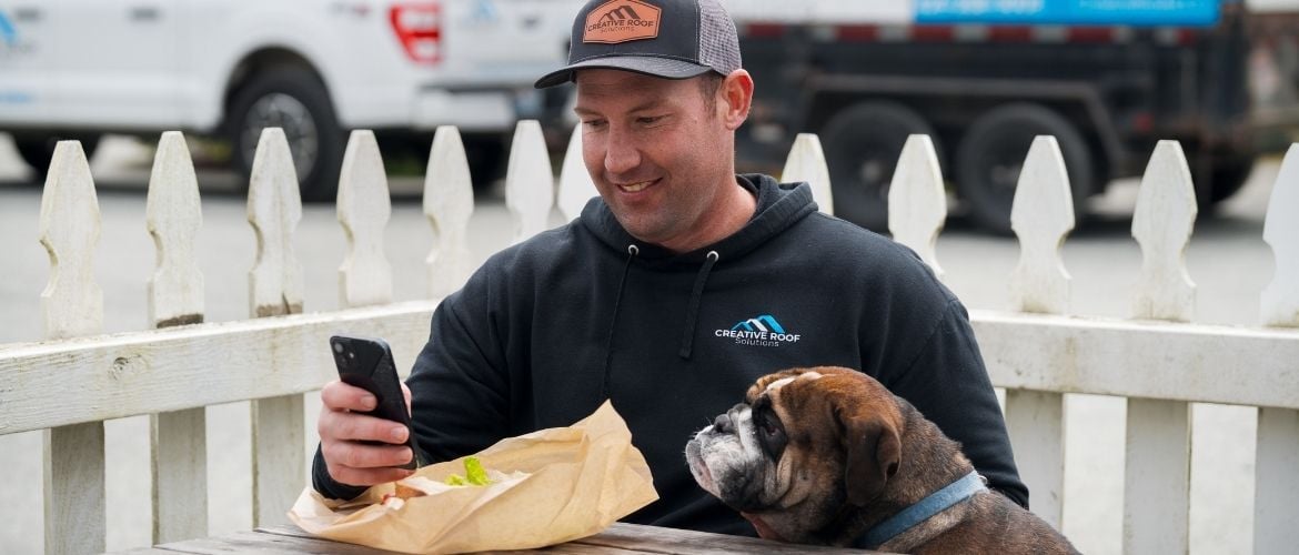 Roofing service provider sits at a table with a bulldog beside him, looking at his phone while eating lunch