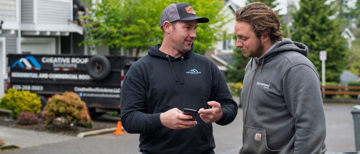 Two roofing service providers stand outside chatting and looking at a smartphone