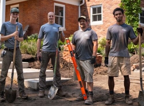 Four landscaping service providers with tools standing in front of a brick house under renovation