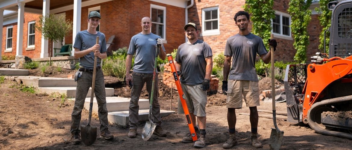 Four landscaping service providers with tools standing in front of a brick house under renovation