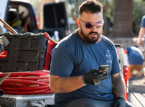 Construction service provider sits on the back of a work truck, checking his phone while other crew members work in the background