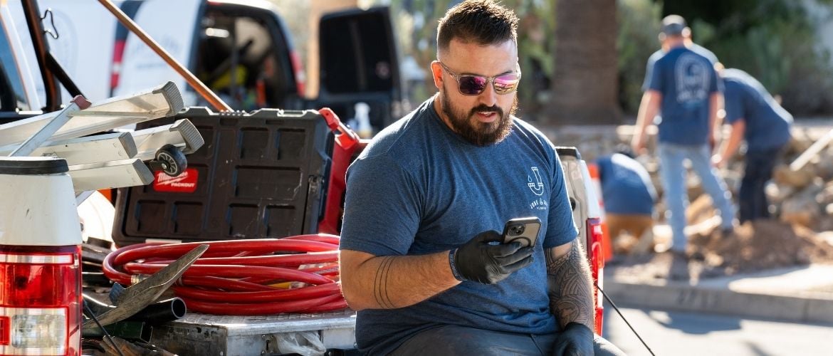 Construction service provider sits on the back of a work truck, checking his phone while other crew members work in the background