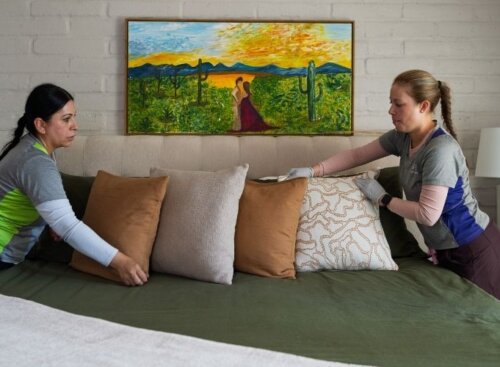Two house cleaners make a bed together in a bedroom with a painting above the headboard