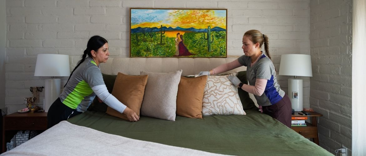 Two house cleaners make a bed together in a bedroom with a painting above the headboard