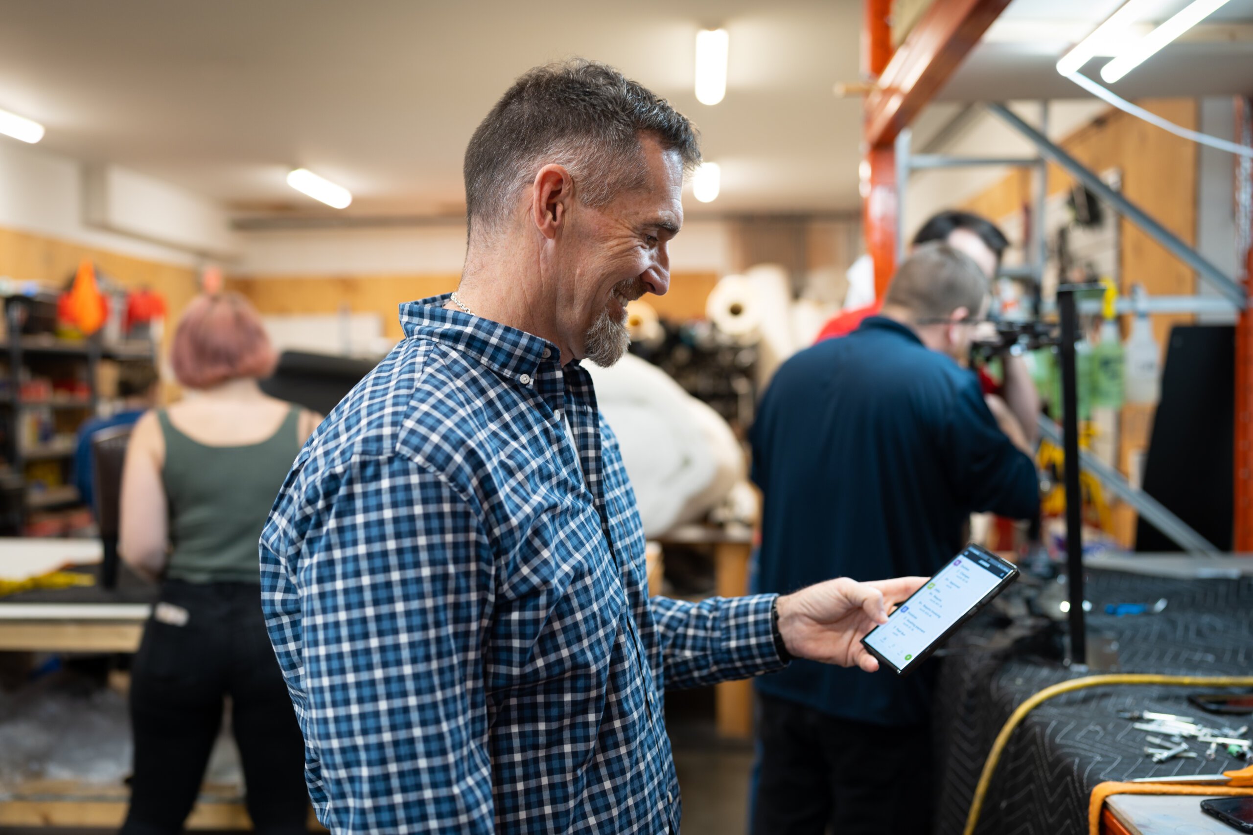 Image shows a franchise owner in a busy leather repair workshop, checking their phone while employees work on pieces in the background.