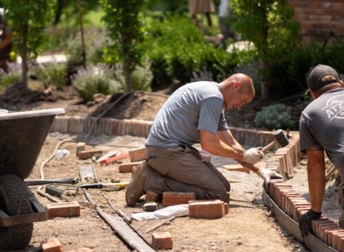 Two landscapers work together to lay bricks along the outside of a garden bed.