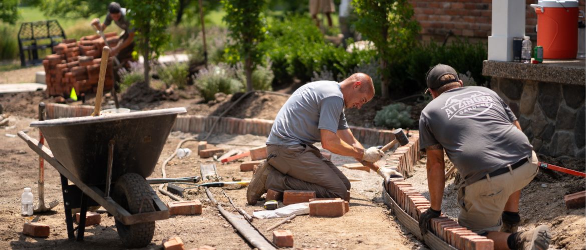Two landscapers work together to lay bricks along the outside of a garden bed.