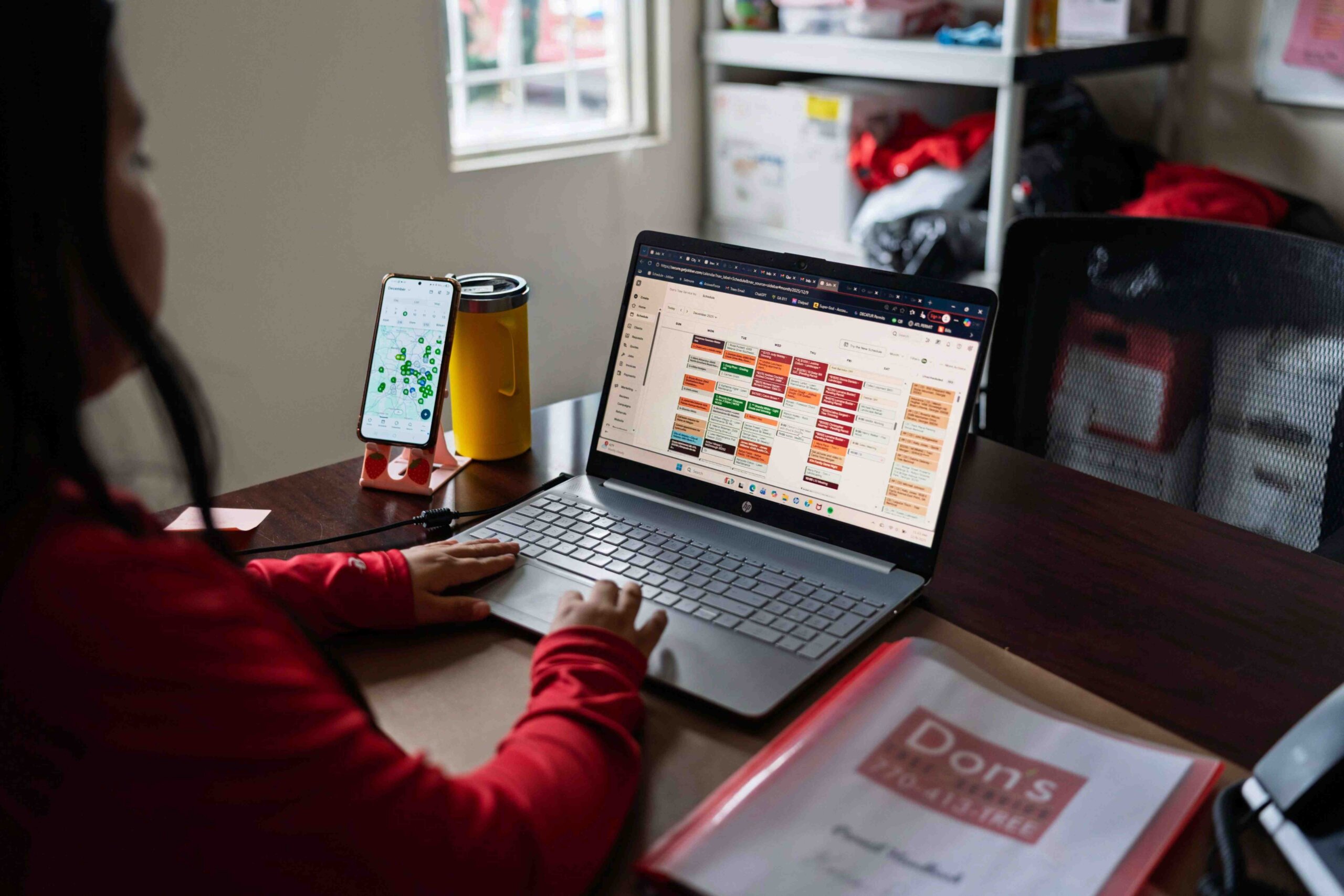 Office staff scheduling jobs on a laptop with a calendar view while tracking crews on a mobile device